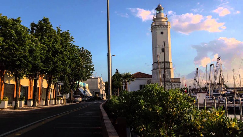 This image shows a street with trees and buildings along it and in the background is a lighthouse on a dock. There are also several bushes and trees scattered throughout the scene