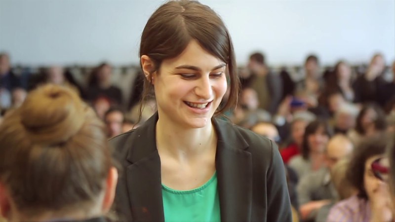 A woman with her hair tied up is wearing a green tshirt with a blazer on top. Blurred in the background there are people sitting and standing up.