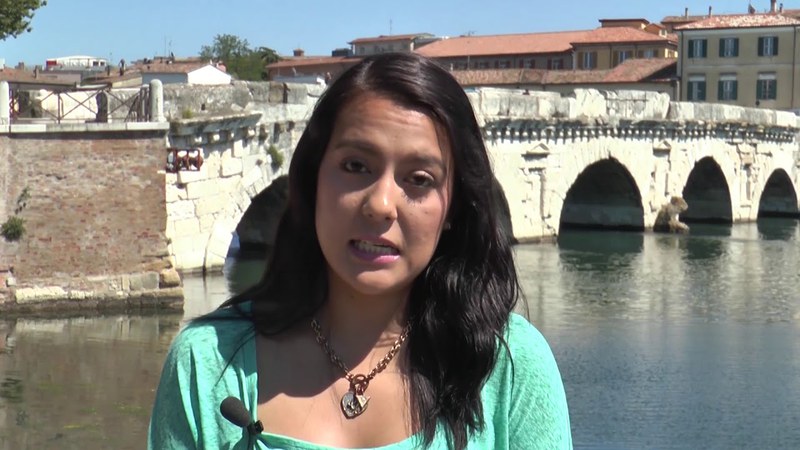 A woman with brown hair is talking to the camera.. She is wearing a teal shirt and a chain necklace. Behind her there is a bridge crossing a river.