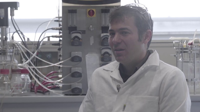 Un uomo con i capelli castani e un camice bianco sta parlando all'interno di un laboratorio. In testa porta degli occhiali protettivi di plastica.