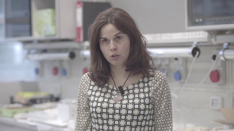 Una donna con i capelli rossi e una maglia a fantasia geometrica parla alla telecamera in un laboratorio.