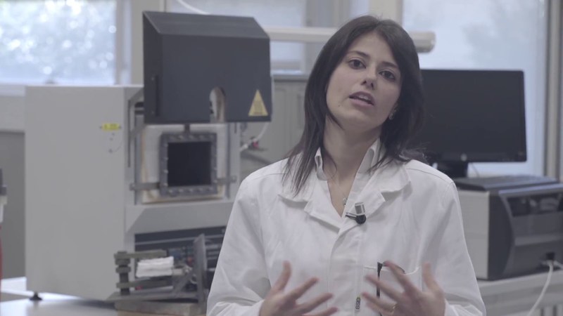 Una donna con i capelli castani che indossa un camice bianco è all'interno di un laboratorio e parla con la telecamera.