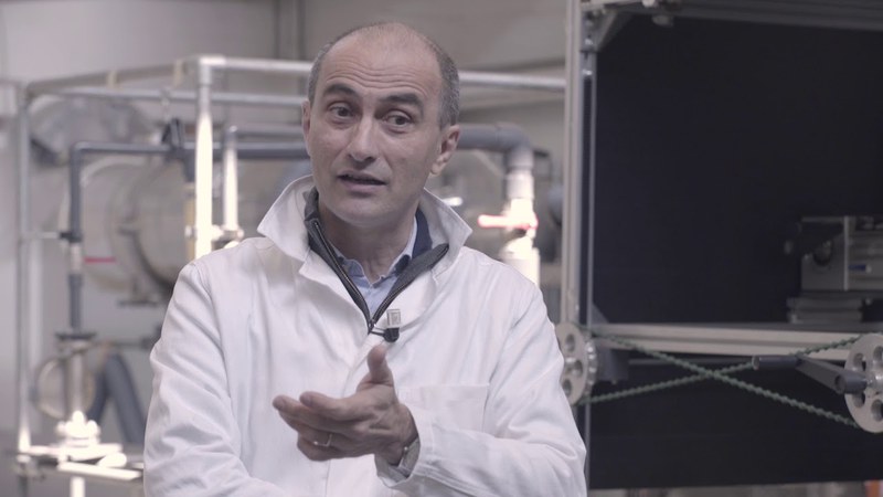 Un uomo con i capelli grigi e un camice bianco sta parlando all'interno di un laboratorio.